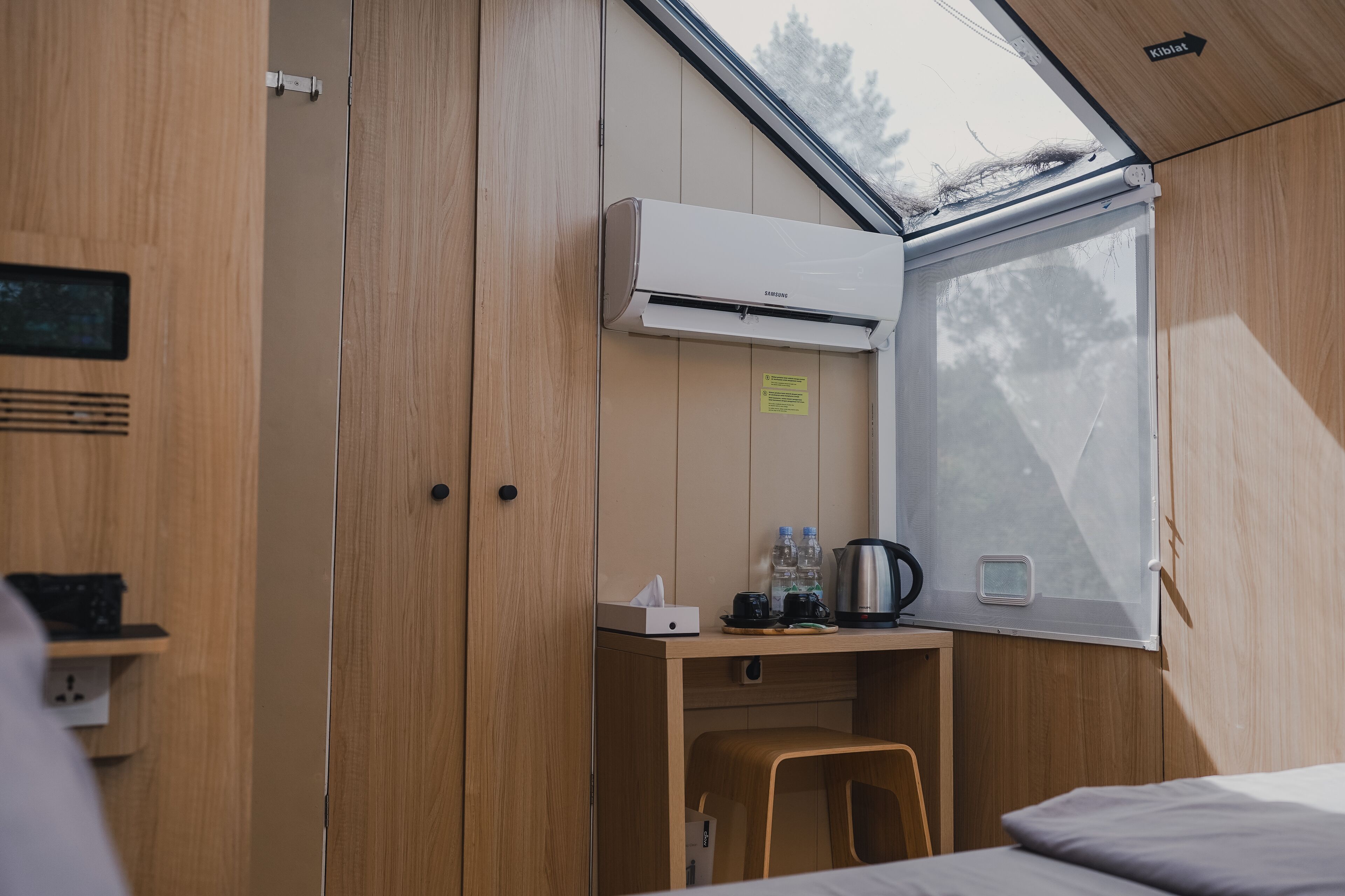Habitación pequeña con interior de madera, un escritorio con un hervidor, una taza y botellas, y una ventana con vistas a árboles.