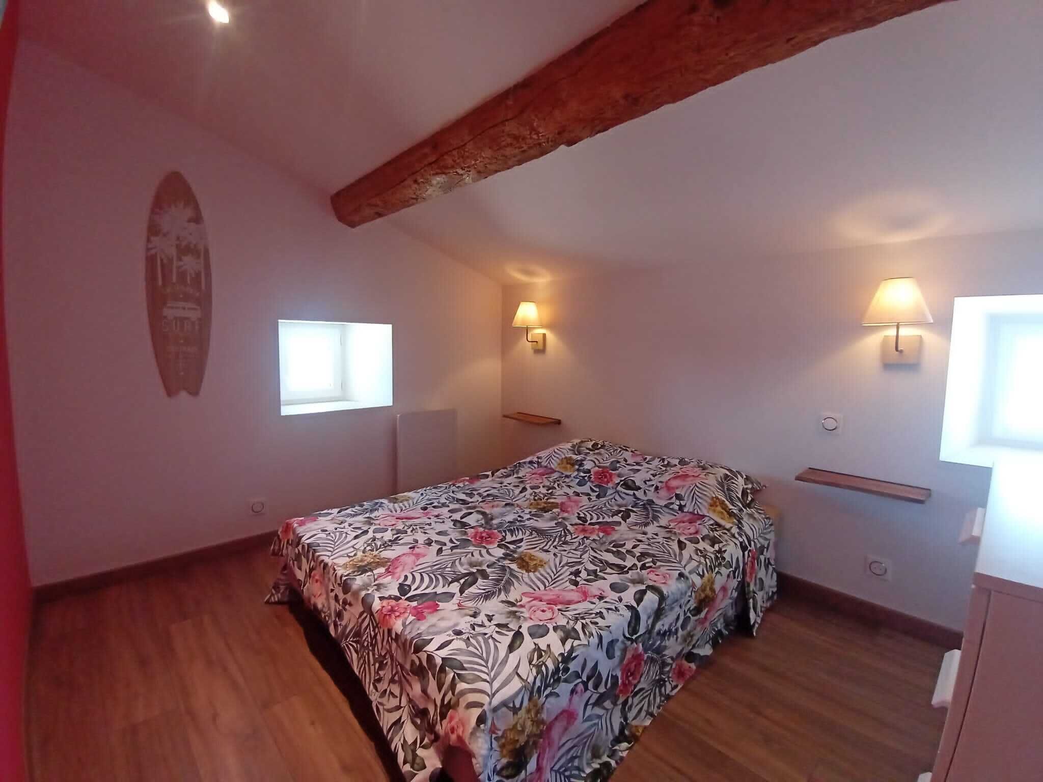 A bedroom with a bed, two wall-mounted lamps, a window, and a wooden ceiling beam.
