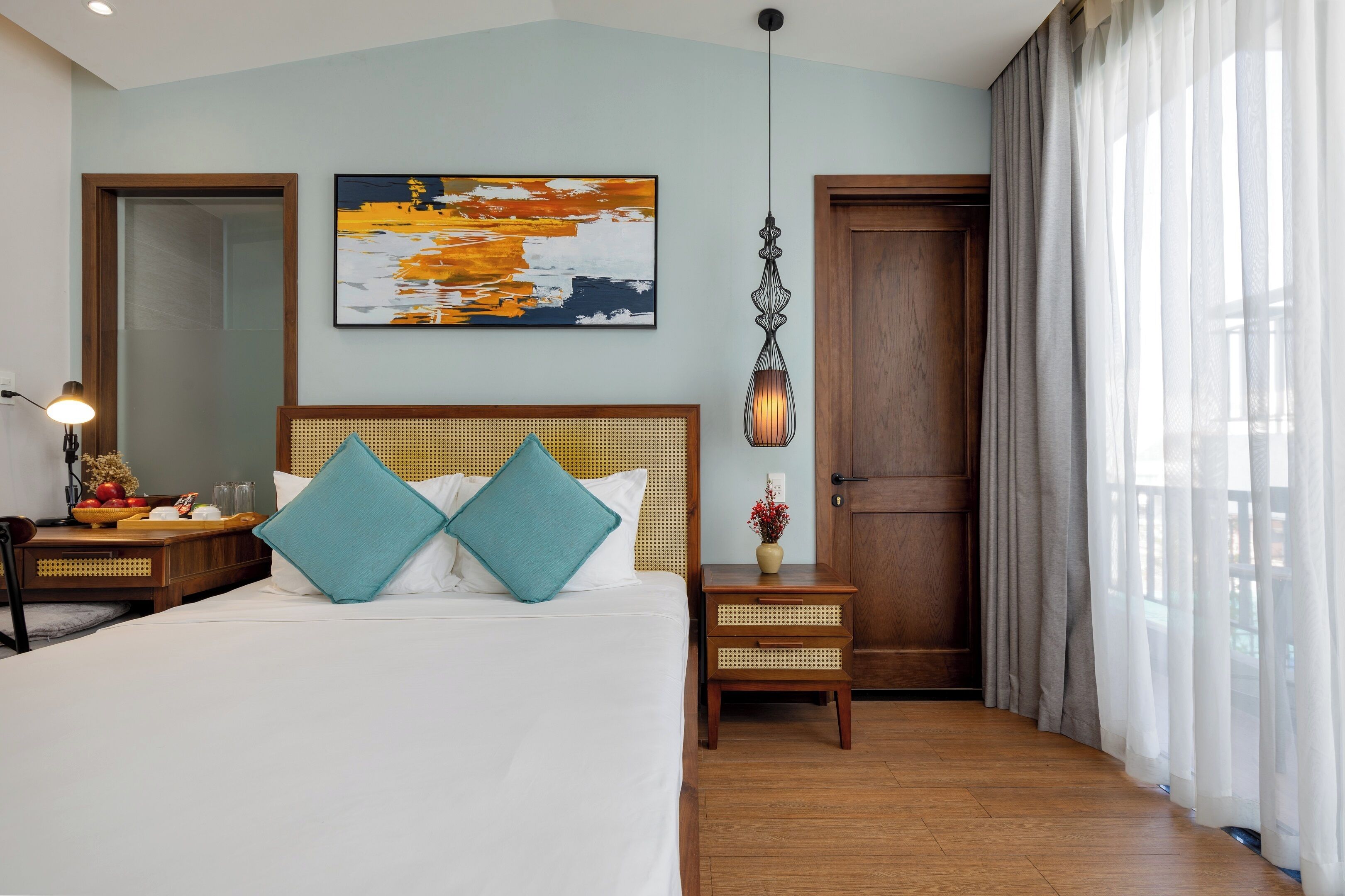 A hotel room with a large bed, a wooden headboard, a nightstand with a lamp, a wall-mounted abstract painting, and a door with curtains.
