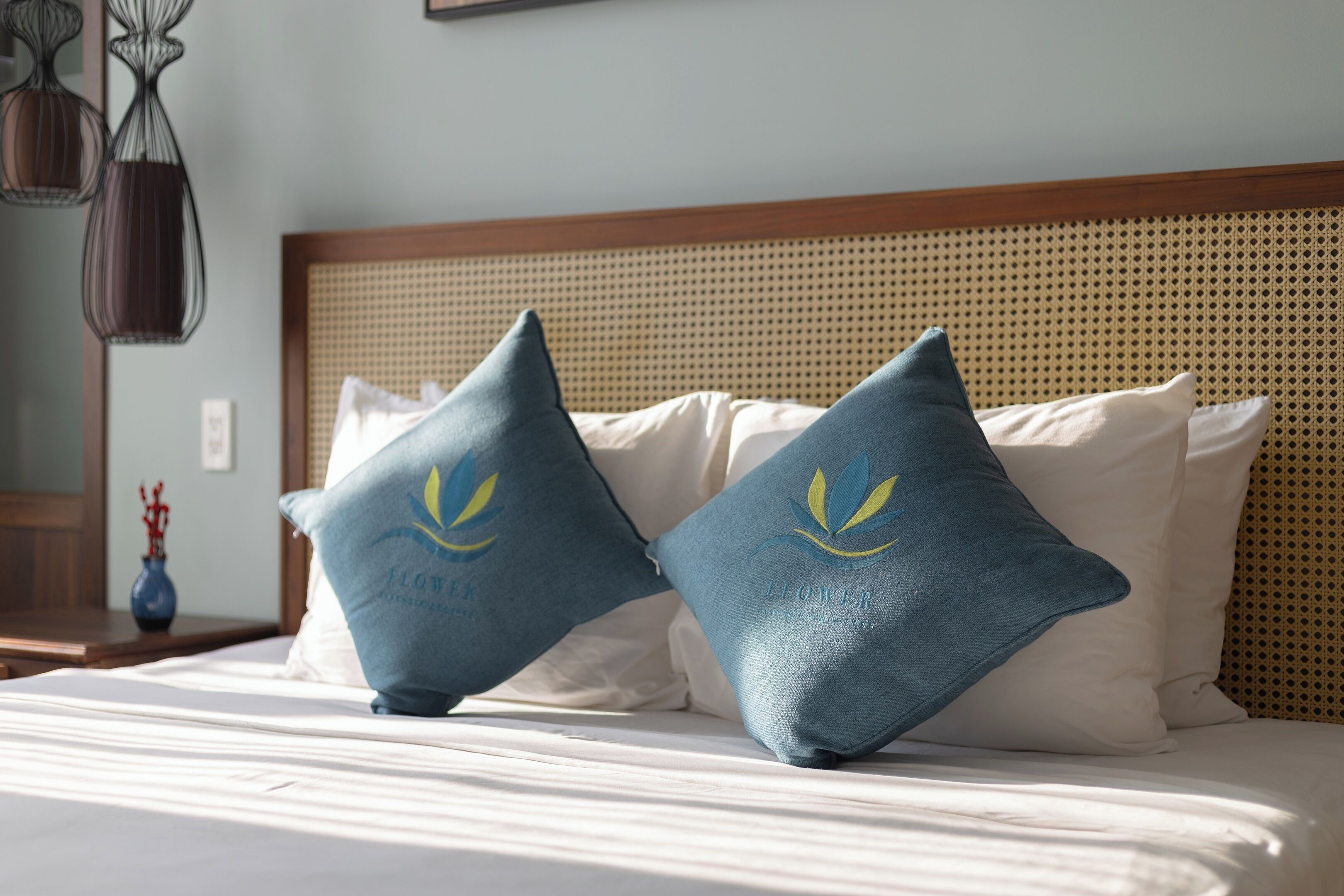 A bed with two pillows featuring a floral logo, a wooden headboard, and a vase on a nightstand.