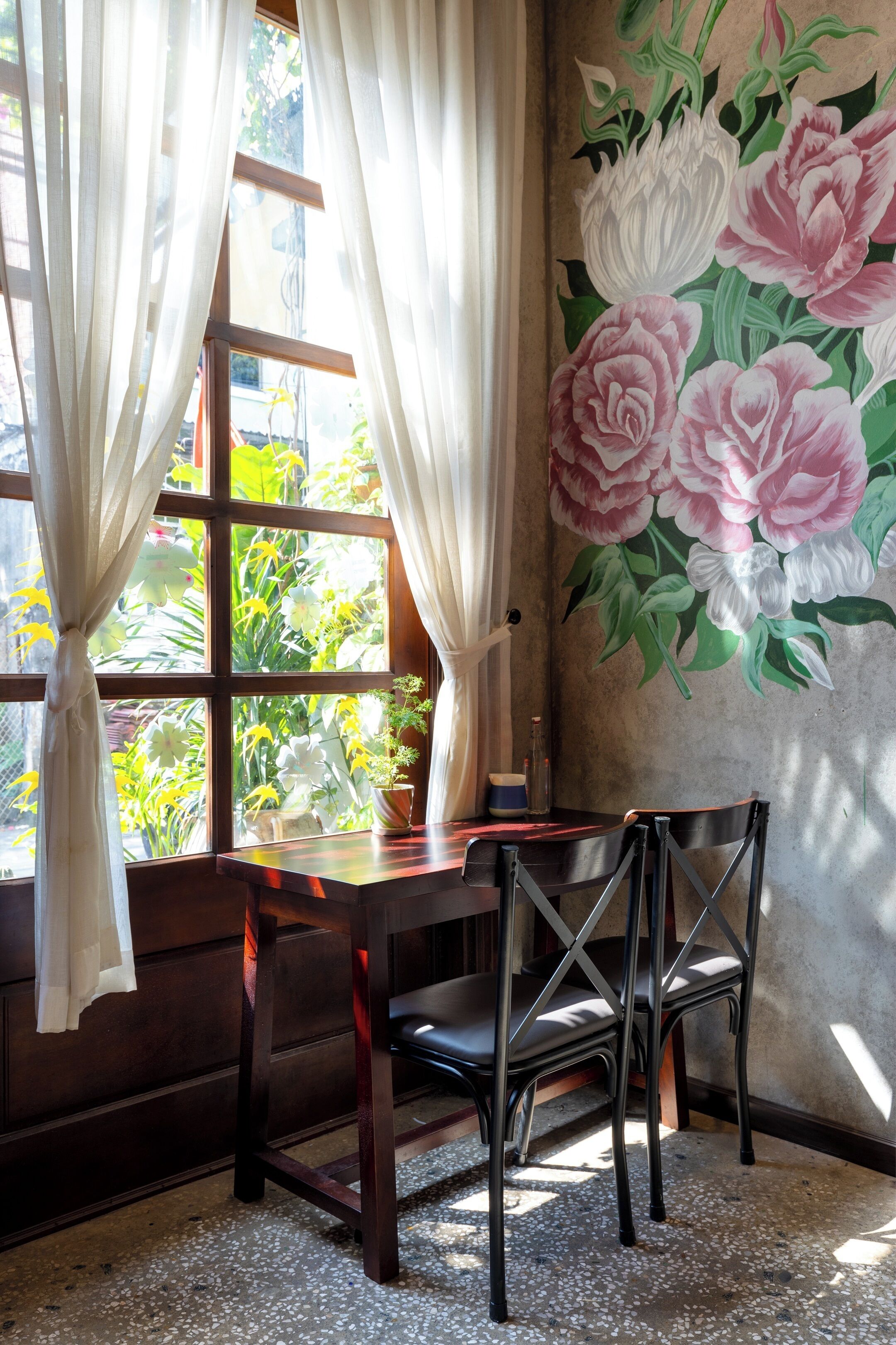 A room with a wooden table and chairs, a window with curtains, and a wall with a floral mural.