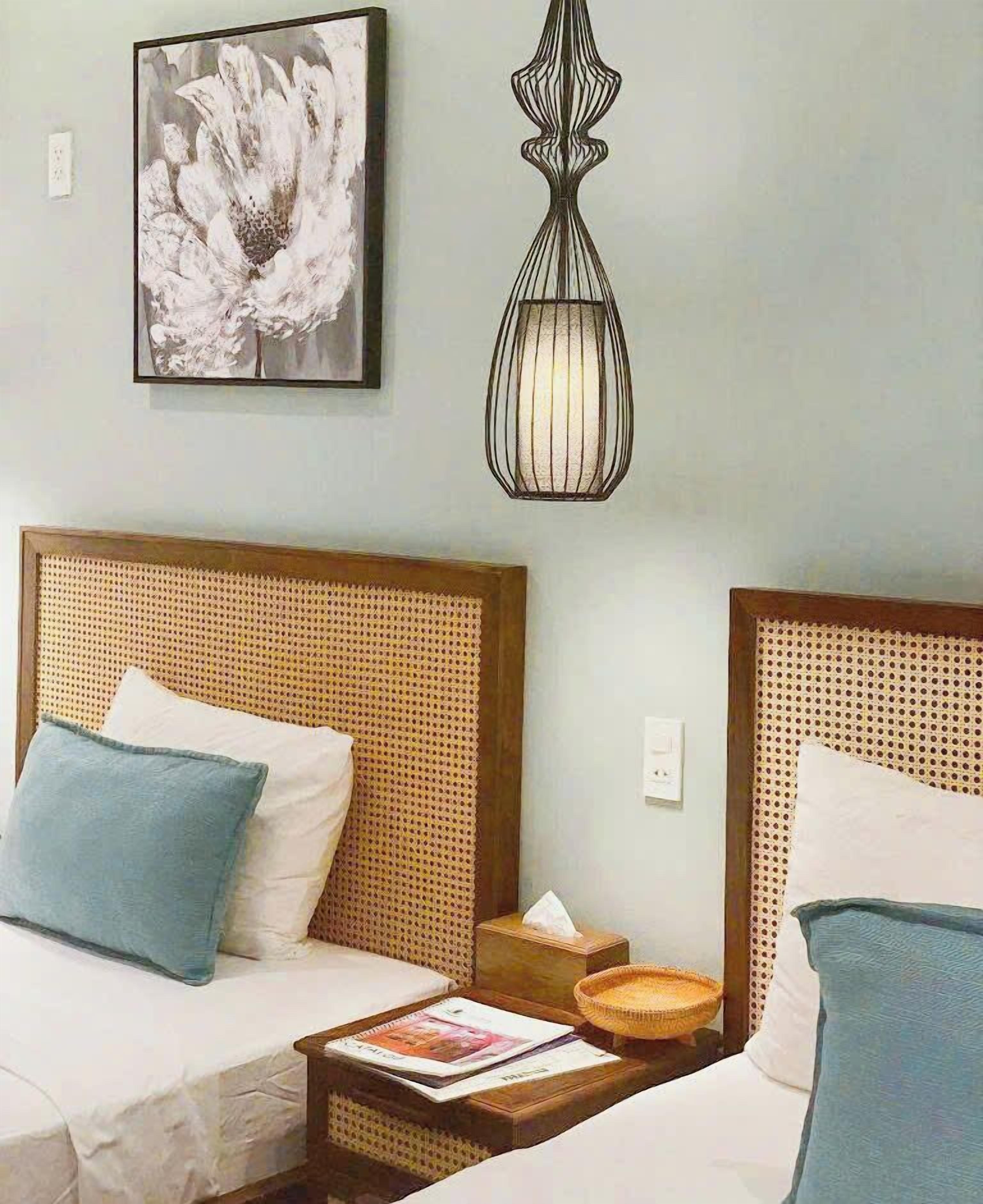 A hotel room with two beds, a rattan headboard, a framed artwork, a hanging light fixture, and a bedside table with a magazine and a tissue box.