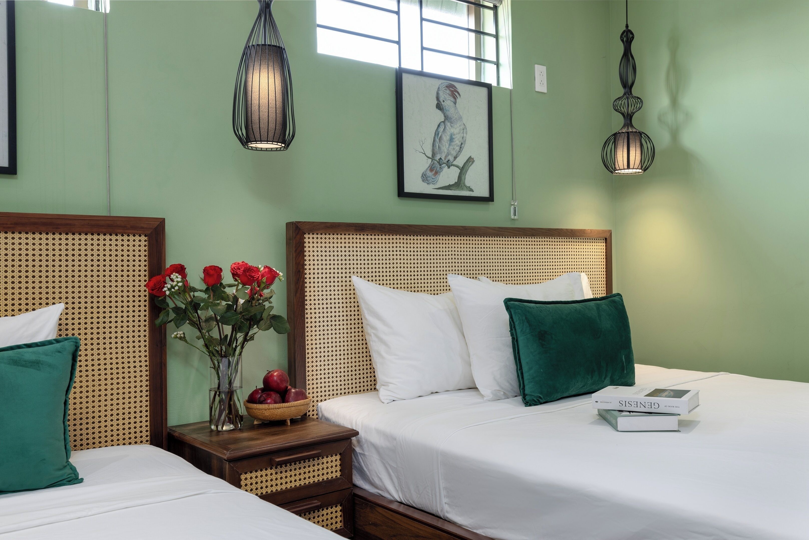 A hotel room with two beds, a rattan headboard, a vase with red flowers, and a framed artwork on the wall.