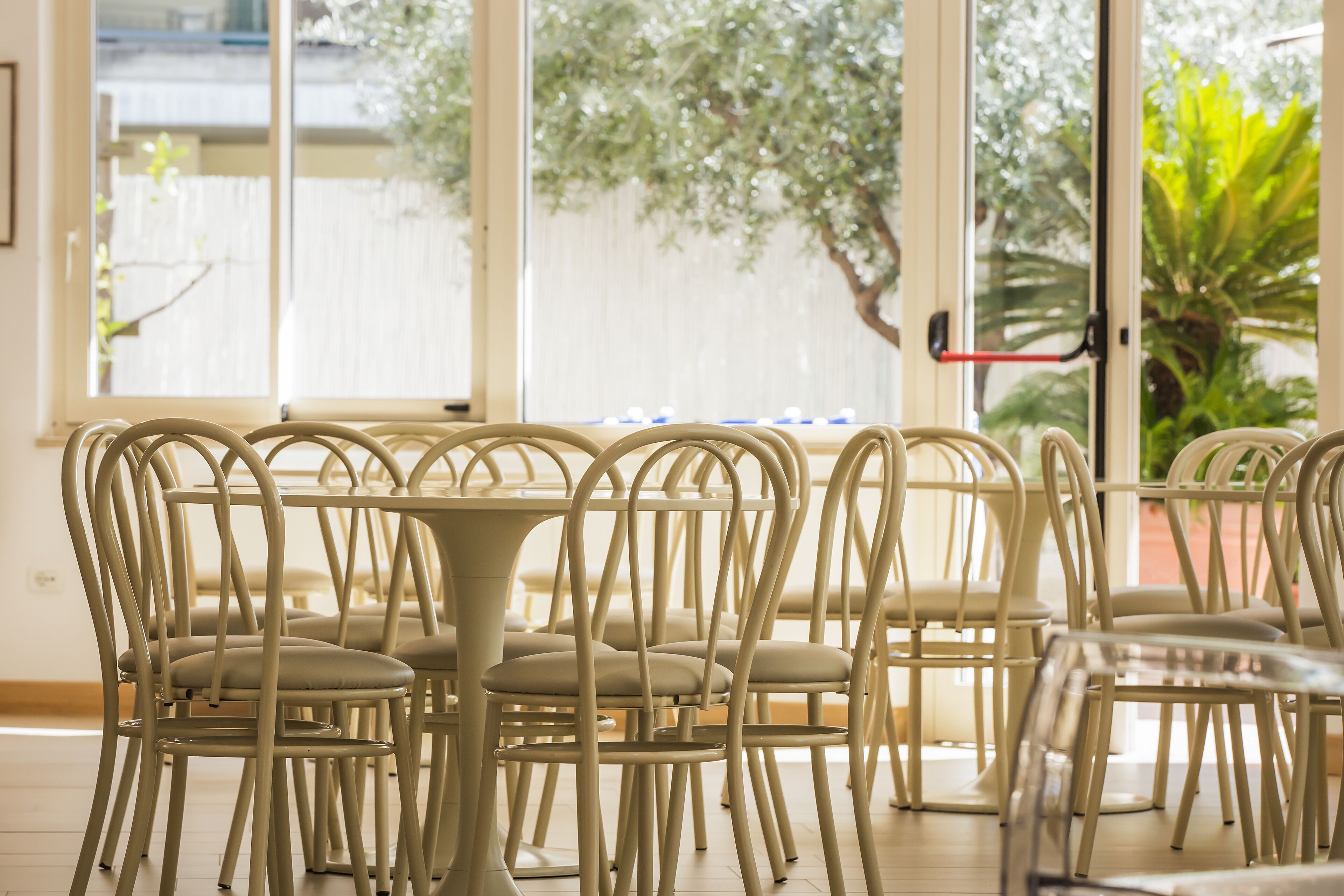 A dining area with white metal chairs and tables, large windows with a view of greenery outside.