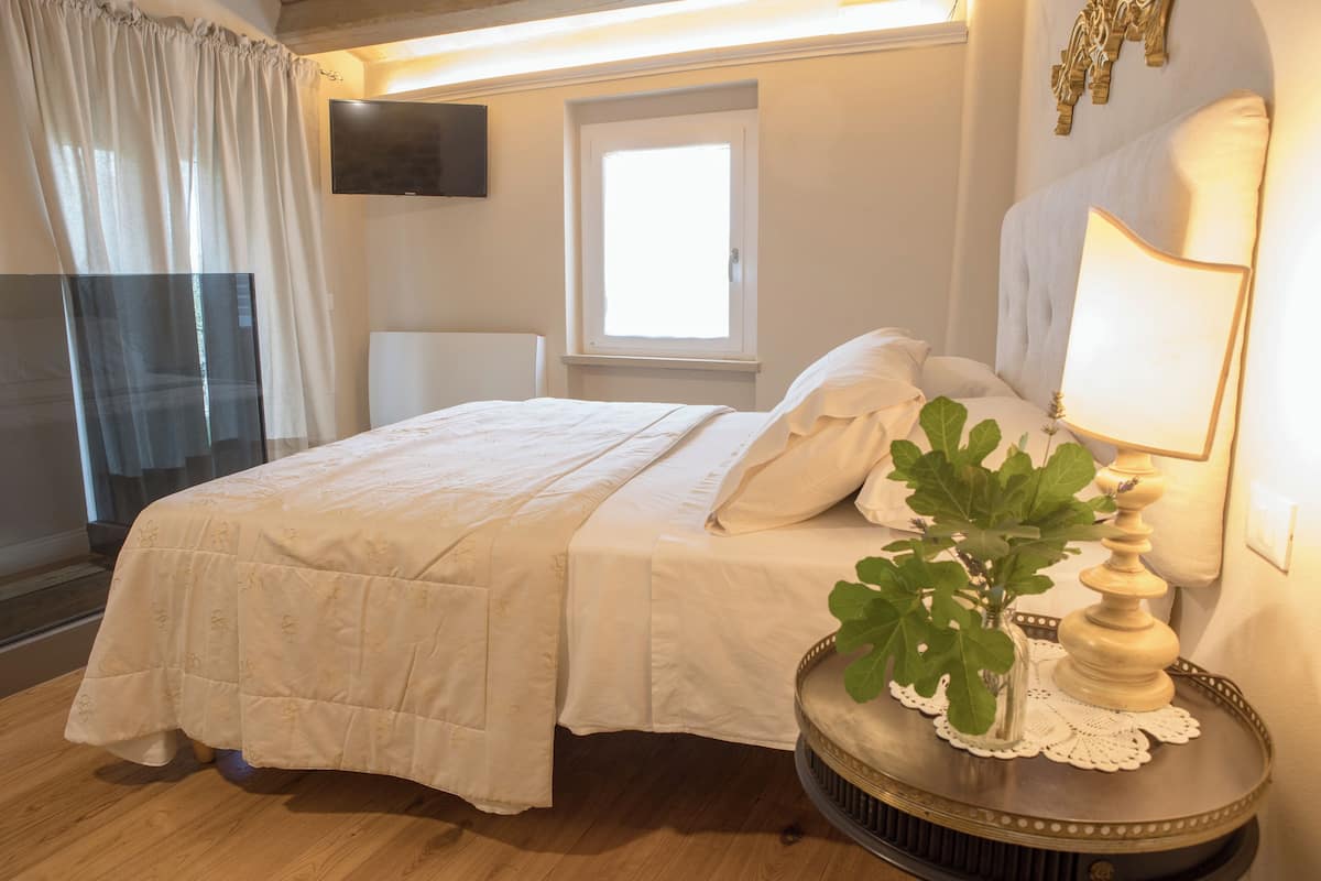 A neatly made bed with a white bedspread and pillows, a bedside table with a lamp and a plant, and a window with curtains.