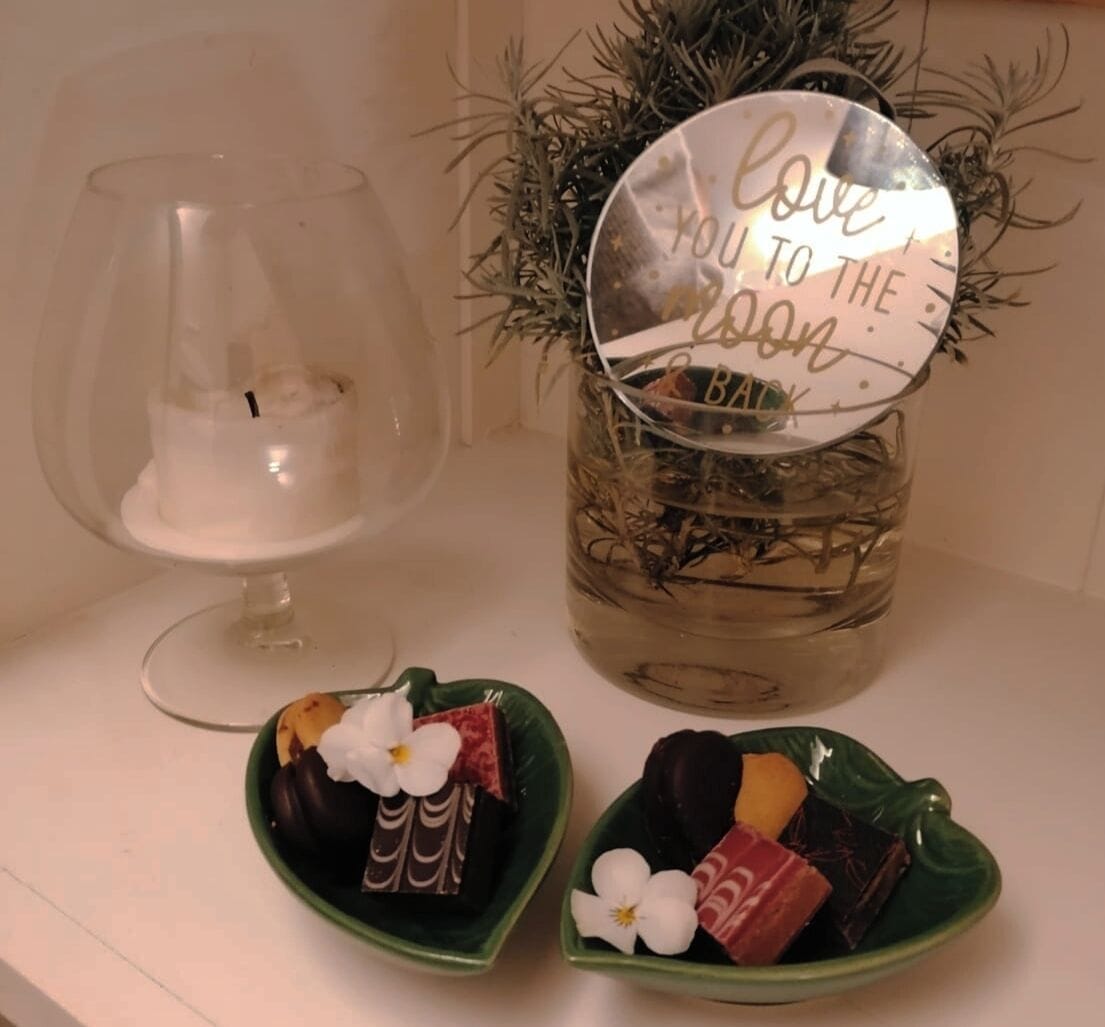 Um castiçal de vidro decorativo, um pote com tampa refletora e duas tigelas em formato de coração com chocolates e flores.