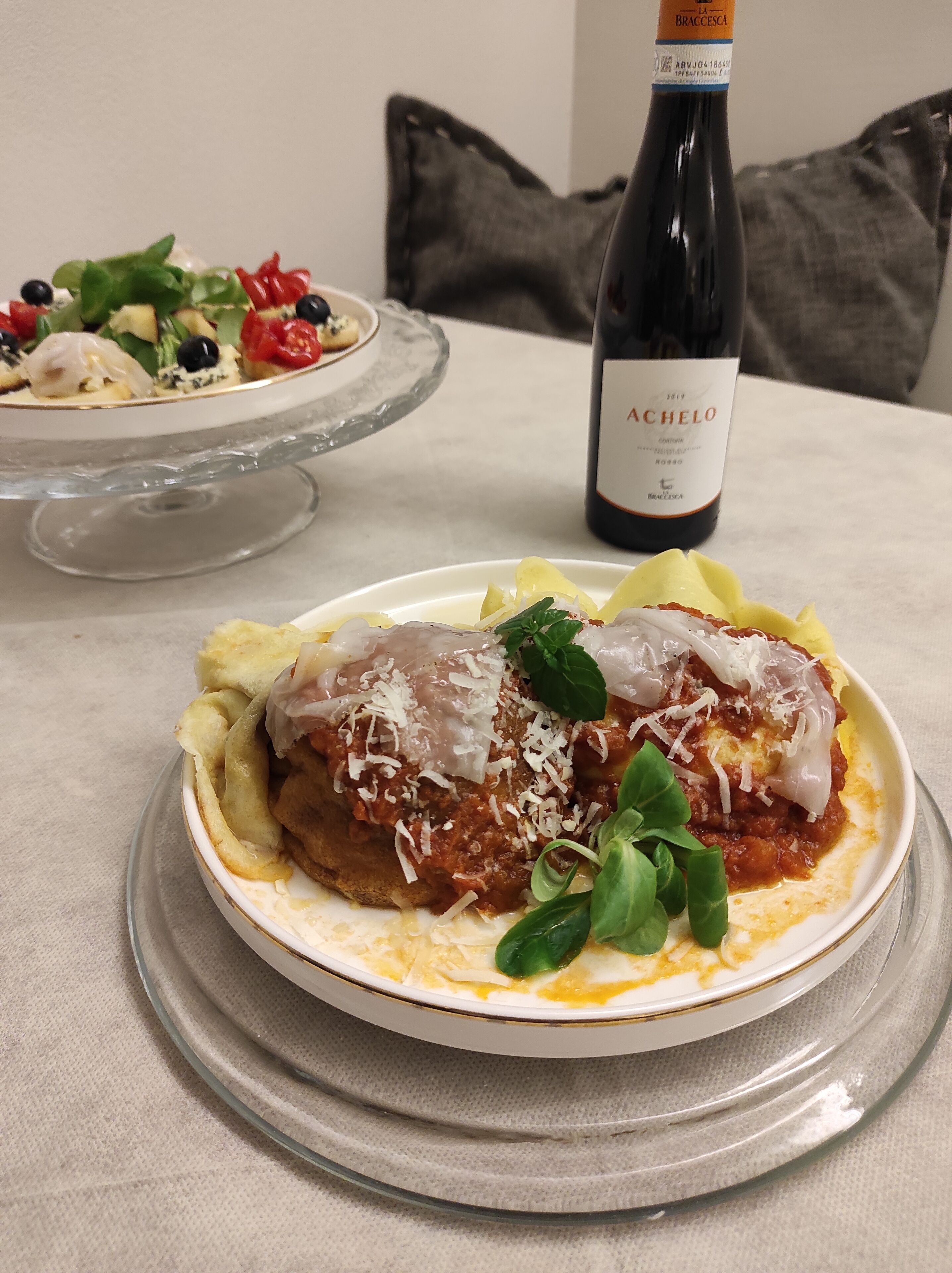 Une assiette de poulet parmesan, avec une bouteille de vin Achelo en arrière-plan.