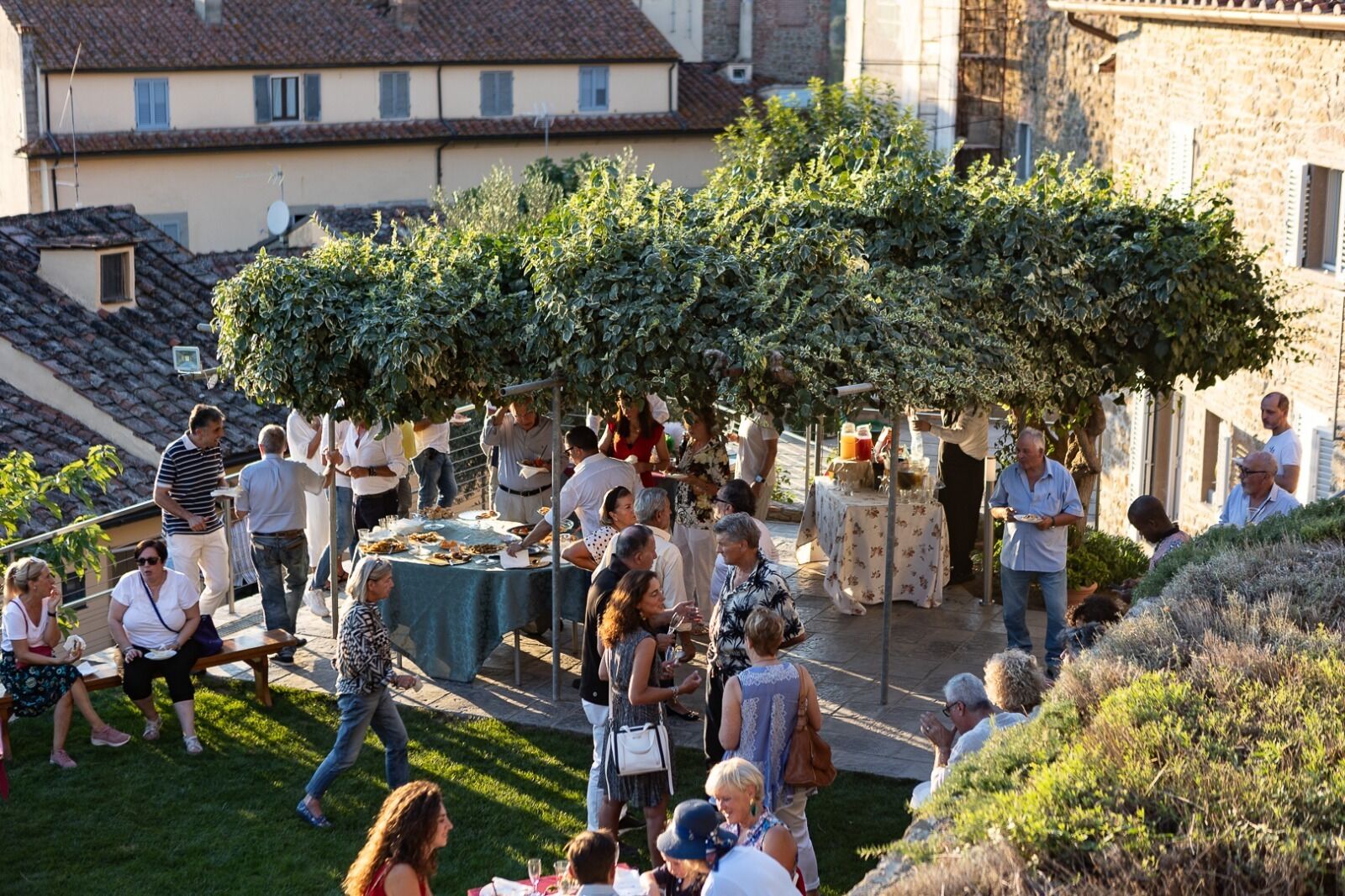An outdoor gathering with people around tables, a grassy area, and buildings in the background.