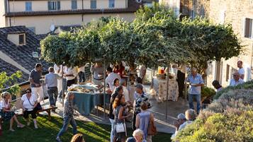 An outdoor gathering with people around tables, a grassy area, and buildings in the background.