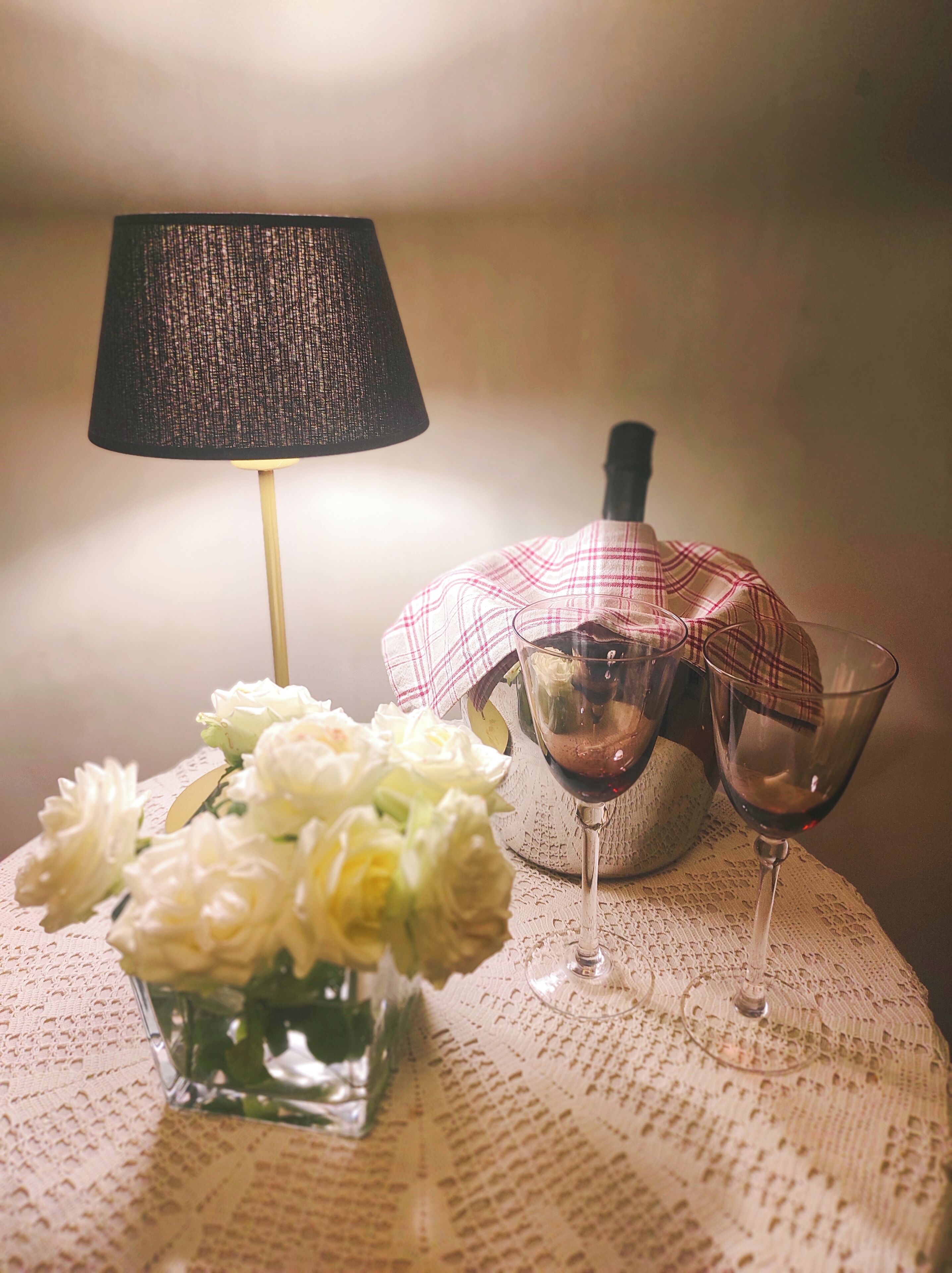 Ein Tisch mit einer Spitzenleinentischdecke, einer Vase mit weißen Blumen, einer Lampe und zwei Weingläsern.