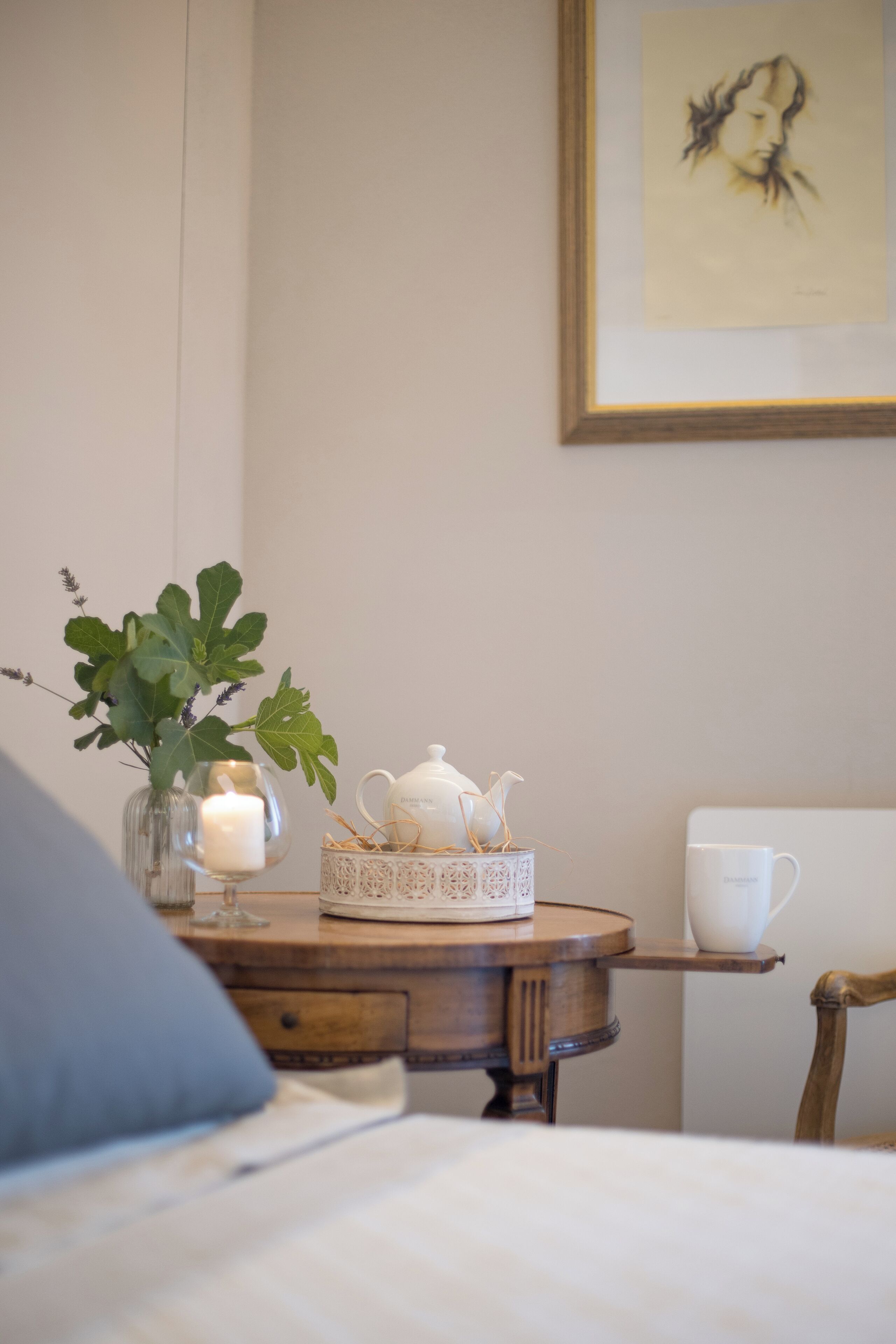 Een knus interieur met een houten tafel, een vaas met groene bladeren, een brandende kaars, een theepot en een kunstwerk in een lijst aan de muur.