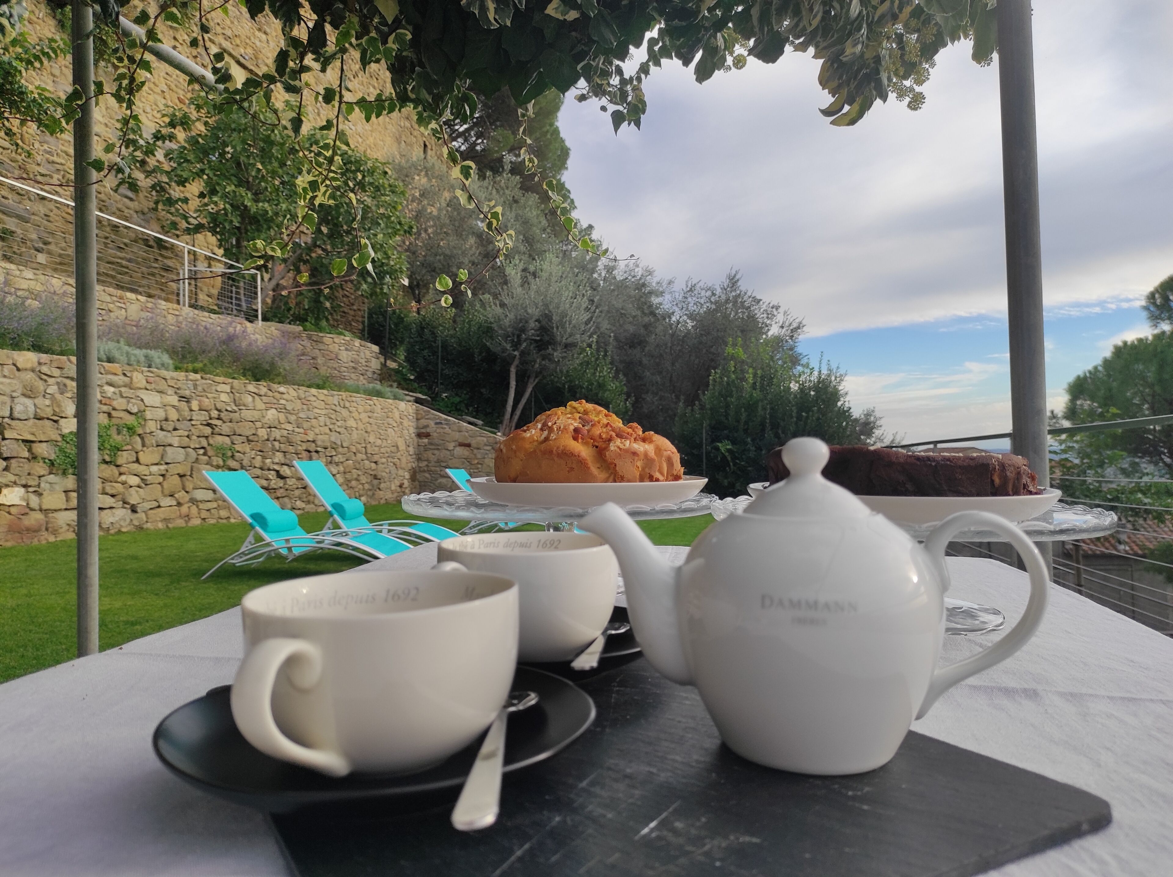 Una mesa con un calentador de agua, tazas y un pastel en una terraza con un muro de piedra y follaje al fondo.