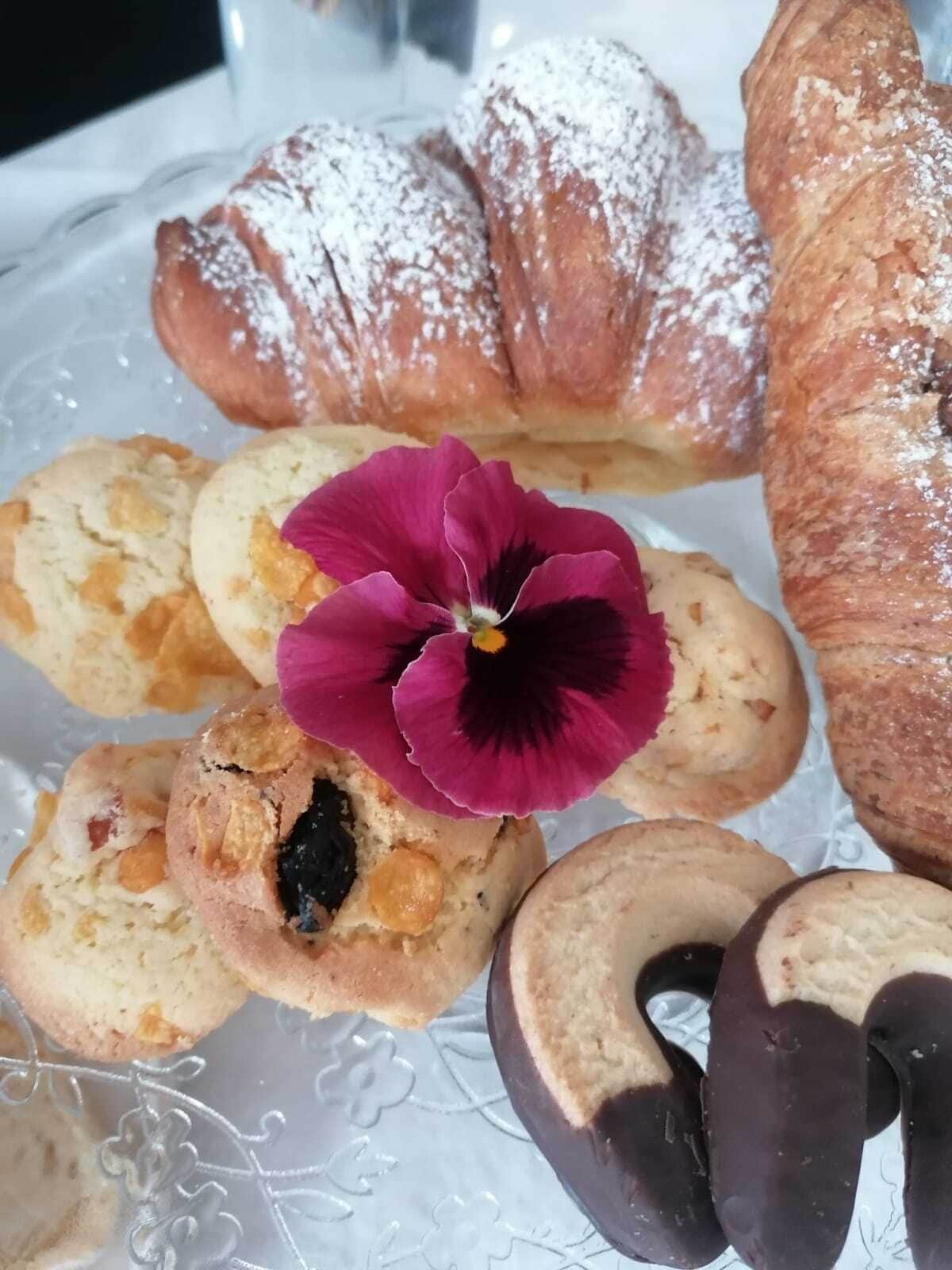 Een bord met gebak, waaronder een croissant, koekjes en chocoladebedekte lekkernijen, met een paarse bloem erbij.