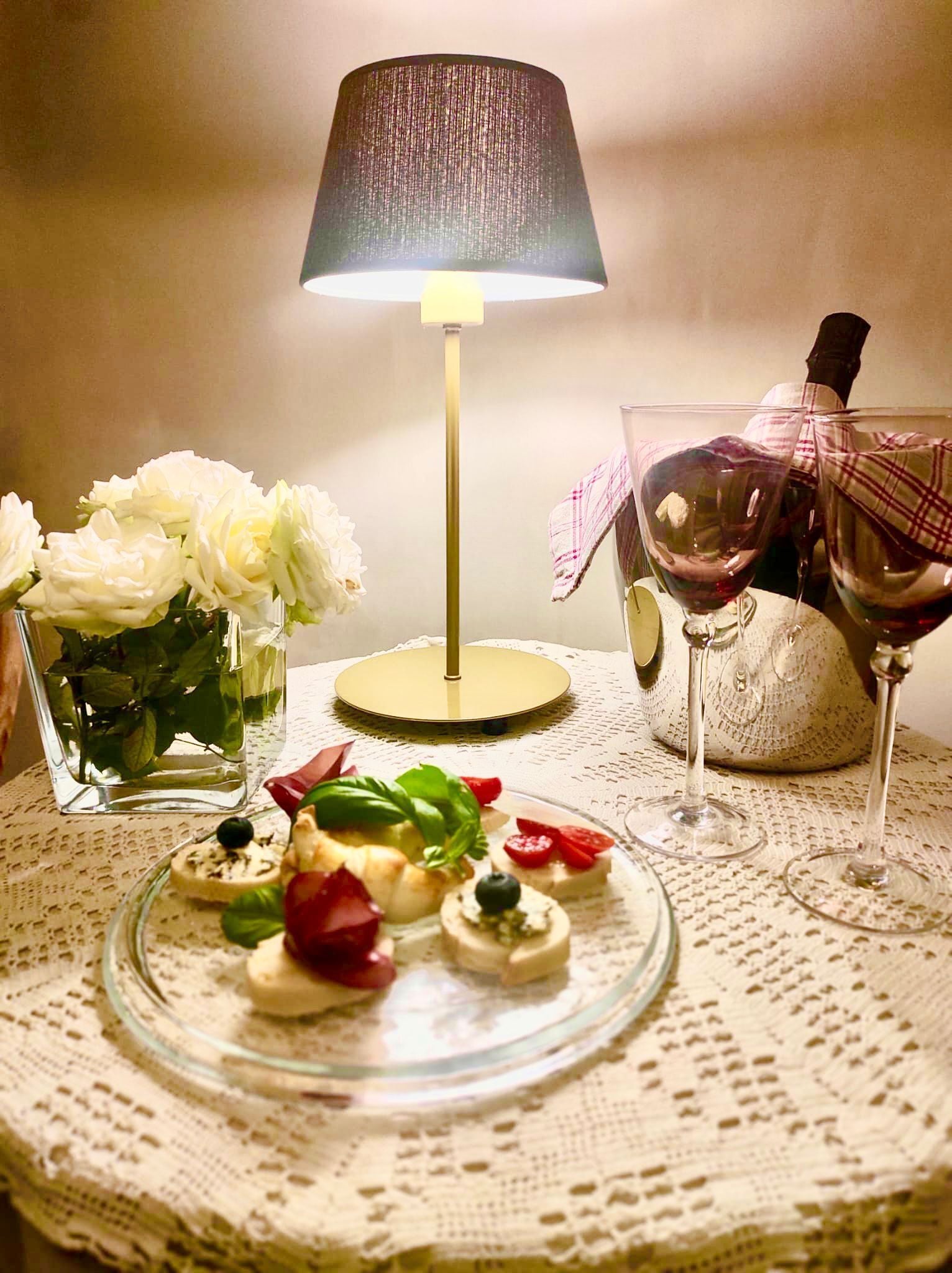 A table with a lace tablecloth, a glass plate with appetizers, a vase with white flowers, a lamp, and two wine glasses.