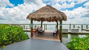 Een houten steiger die uitkomt bij een gazebo met rieten dak en banken, omgeven door weelderige begroeiing en een rustig waterlichaam.