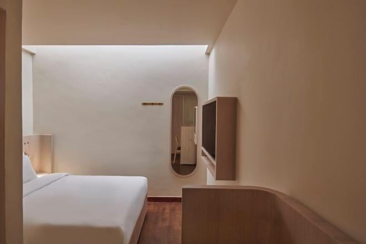 A hotel room with a bed, a small desk, a mirror, and a TV mounted on the wall.