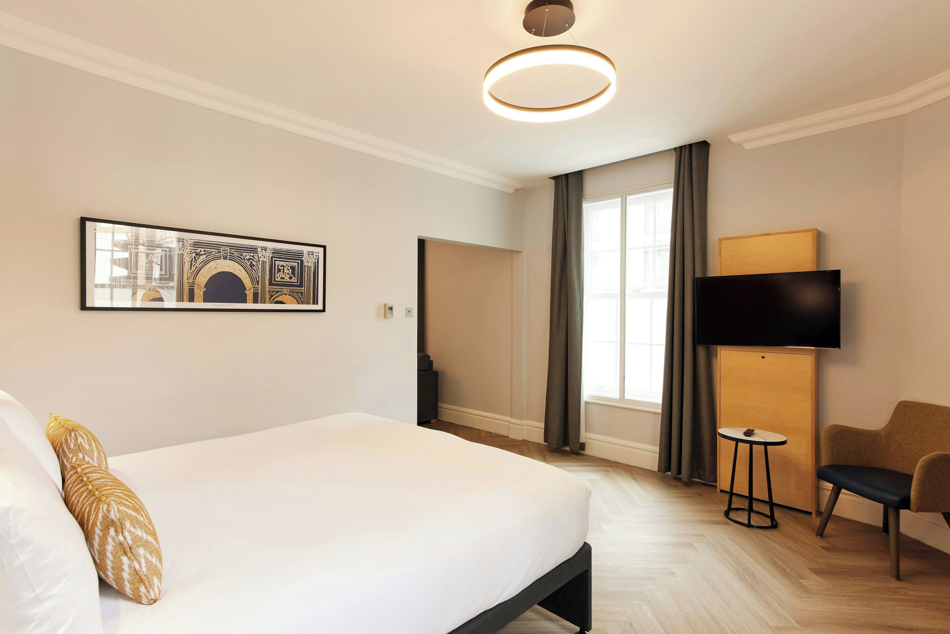 A hotel room with a bed, a flat-screen TV, a small table, a chair, and a painting on the wall.