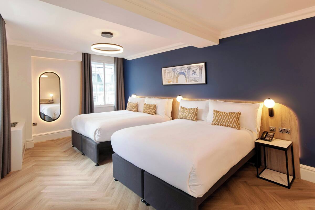 A hotel room with two beds, a mirror, a bedside table with a phone, and a painting on the wall.