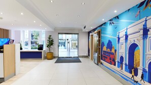 A modern hotel lobby with a reception desk, a large potted plant, and a mural of a cityscape on the wall. - Thistle London Park Lane (London)