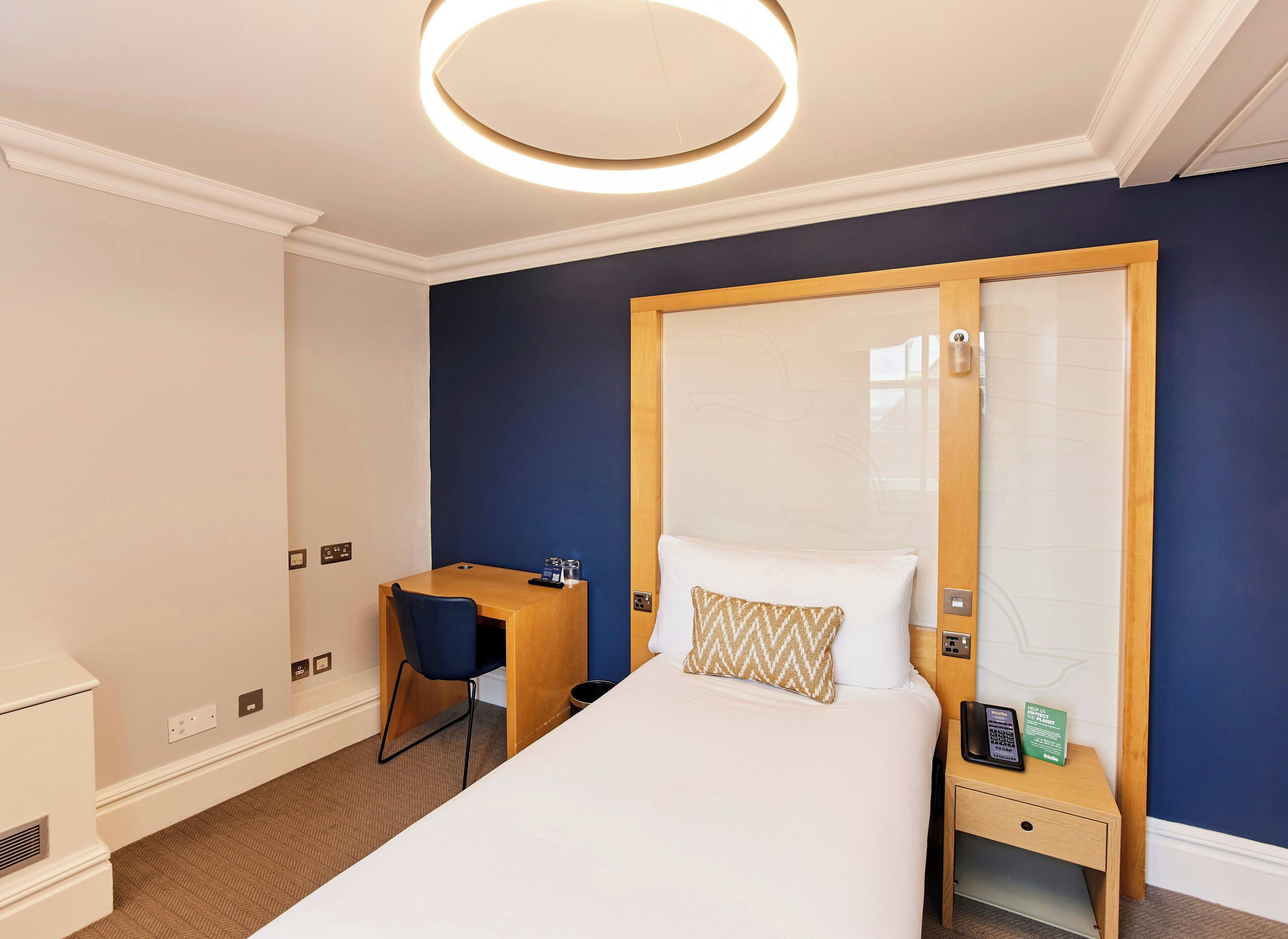 A hotel room with a bed, a desk, a chair, a nightstand, a phone, and a wall-mounted light fixture.