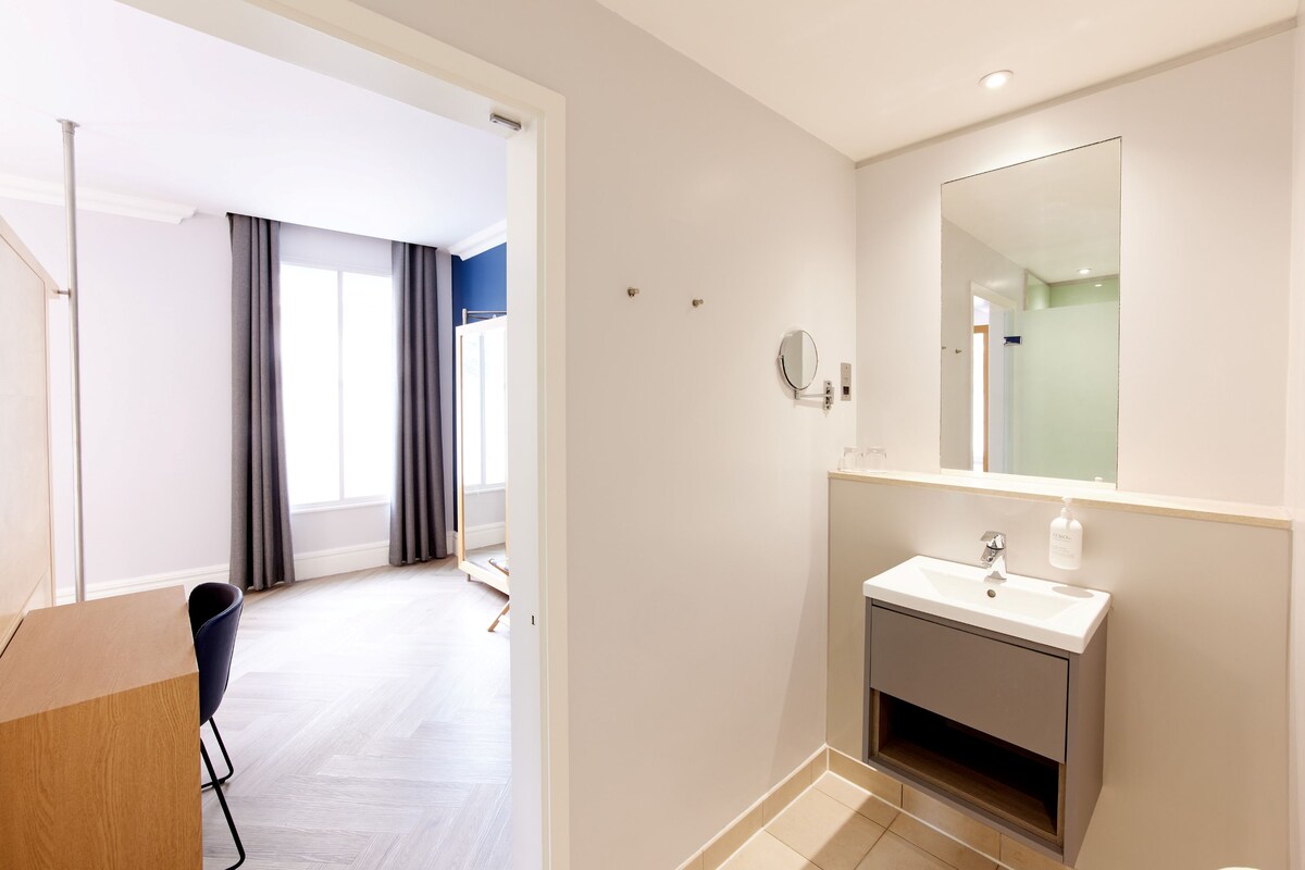 A hotel room with a bathroom, a wooden desk, a chair, a bed, and a view of the hallway.