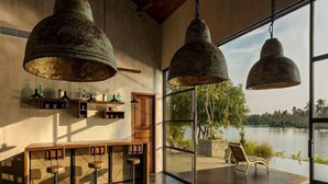 Et moderne interiør med et stort spisebord, hængende lamper i industriel stil og udsigt til en sø gennem store vinduer.
