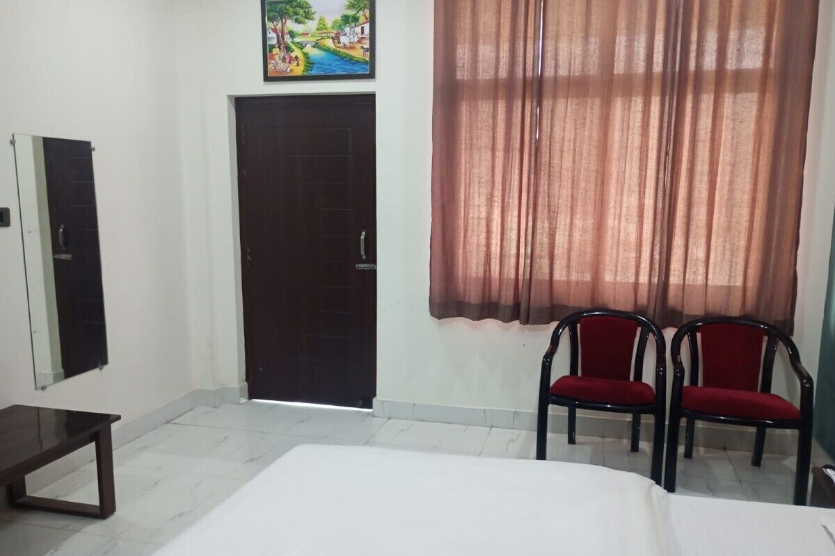 Una habitación con una cama, un banco, dos sillas rojas, una puerta y un cuadro en la pared.