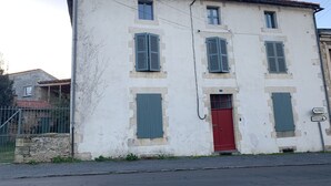 Een gebouw met twee verdiepingen, met een rode deur en groene luiken, een stenen muur en een bordje dat een kampeerterrein aangeeft.