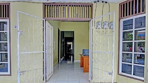 An open gate leading to a hallway with a tiled floor, a wooden desk, and a window with a view of the outside. - Hotel O Firza Homestay (Palembang)