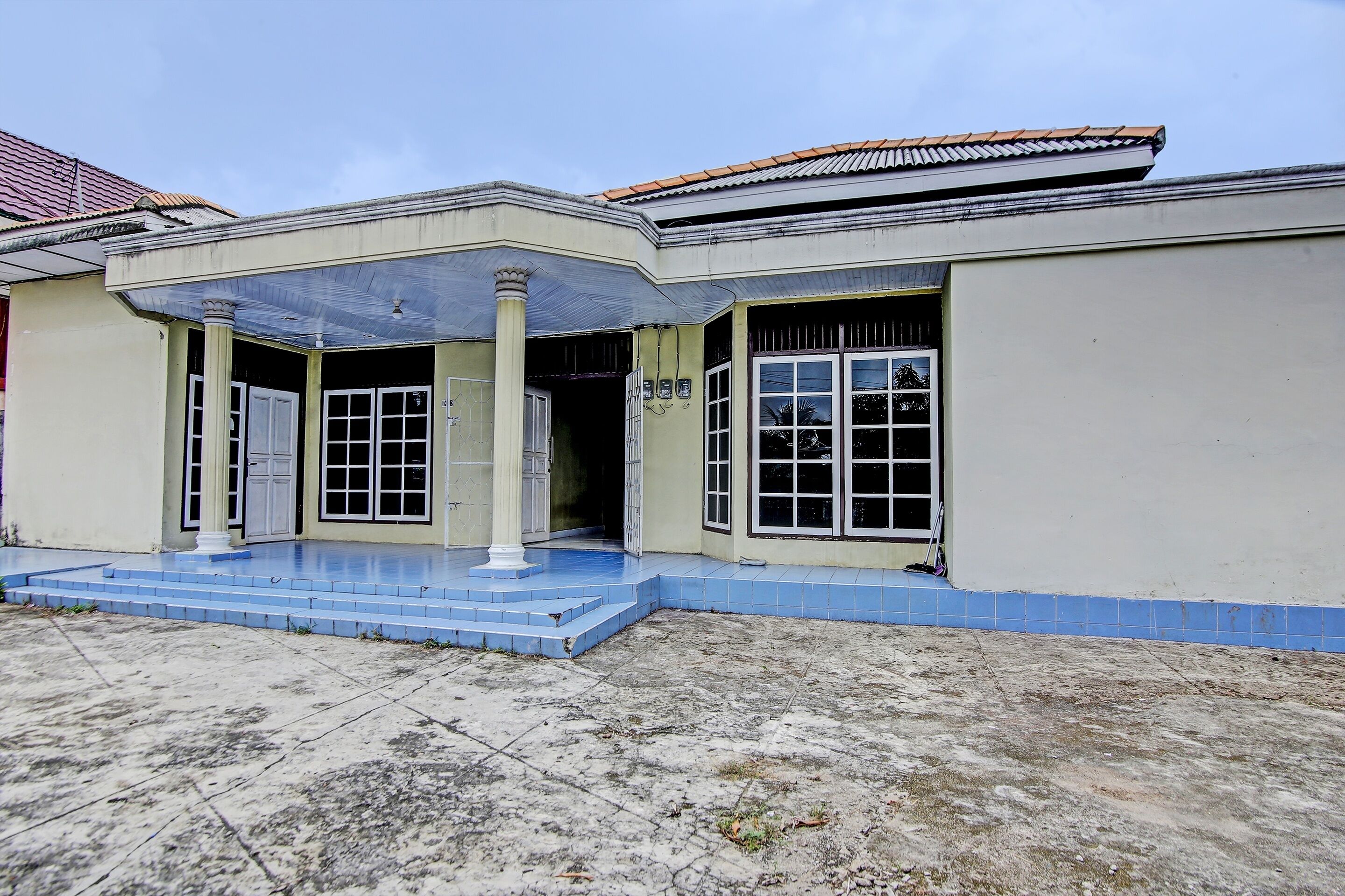 A two-story building with a flat roof, a porch with blue-tiled steps, and a large window with a metal grill.