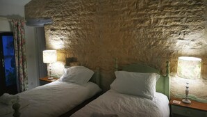 Una habitación con dos camas, cada una con cabecero de madera y una lámpara sobre una mesita de noche. Las paredes tienen textura de piedra.