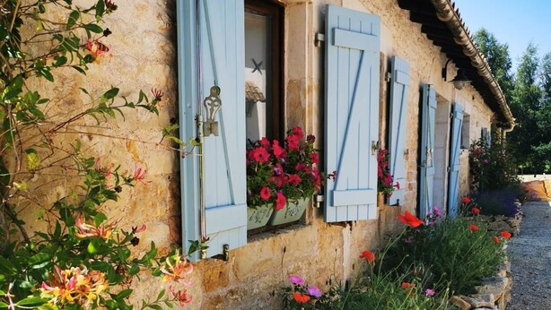 Un edificio rústico de piedra con contraventanas azules, un camino de grava y diversas plantas en flor.