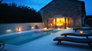 Un área de piscina al aire libre con sillas de descanso y una zona de estar cubierta, rodeada de muros de piedra y árboles.