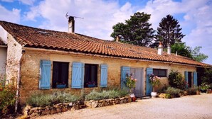 Un edificio de piedra rústico con contraventanas azules, un camino de grava y un jardín con diversas plantas y flores.