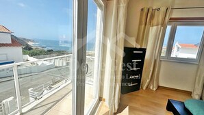 Ein Zimmer mit einem großen Fenster, das Meer und Gebäude zeigt. An der Wand hängt ein schwarzes Schild und in der Ecke steht eine blaue Bank.
