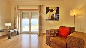Ein geräumiges Wohnzimmer mit einem großen Schiebeglasfenster, das auf einen Balkon führt, ein modernes Sofa mit rotem Kissen, eine Stehlampe und ein minimalistisches Kunstwerk an der Wand.