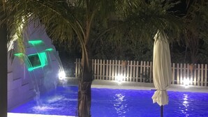 Uma área de piscina à noite, com fontes iluminadas e uma cerca branca ao fundo.