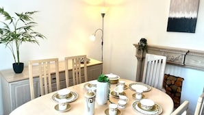 A dining area with a round table set for four, surrounded by chairs. There is a potted plant on the left, a wall-mounted lamp above the table, and a fireplace with a mantel to the right. - Spacious Family Home (Manchester)