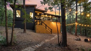 Una moderna baita con una scala in legno che conduce a un patio, circondata da alberi e un sentiero in pietra.