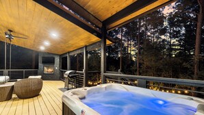 Una vasca idromassaggio su un pontile di legno, con mobili in vimini, un camino in pietra e un'ampia finestra che si affaccia su una foresta.
