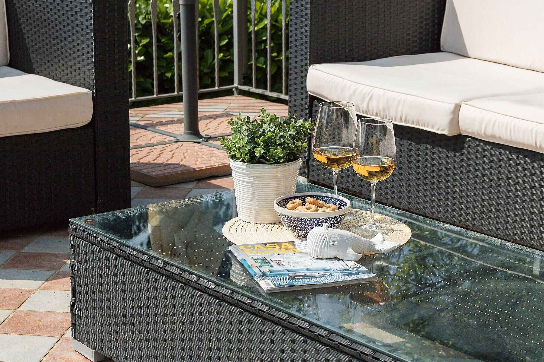 Ein Tisch mit Glasplatte, eine Zeitschrift, eine Schale mit Snacks, zwei Weingläser und ein Blumentopf auf einer Terrasse mit Korbmöbeln.
