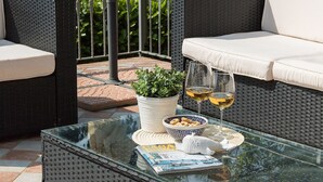 Mesa con cubierta de vidrio, revista, un cuenco de aperitivos, dos copas de vino y una planta en maceta en un patio con mobiliario de mimbre.