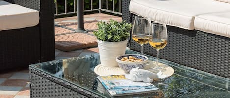 A glass-top table with a magazine, a bowl of snacks, two wine glasses, and a potted plant on a patio with wicker furniture.