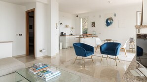 Una sala de estar moderna con una mesa de centro de vidrio, dos sillones azules, un sofá blanco y una cocina al fondo.