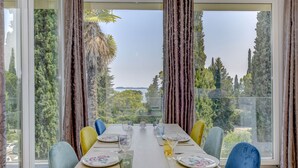 A dining table set for four with floral tableware, positioned in a room with large windows offering a view of trees and a distant landscape. - Apartment Diamante with lake view and pool (Gardone Riviera)