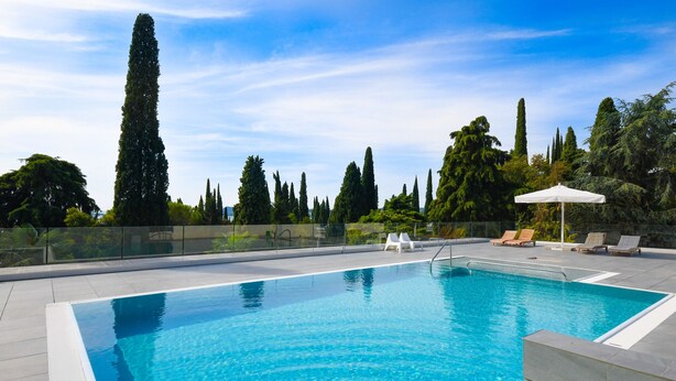 A rectangular swimming pool with clear blue water, surrounded by a tiled deck. There are lounge chairs and a white umbrella on the deck. Tall trees and a clear sky are visible in the background. - Apartment Diamante with lake view and pool (Gardone Riviera)