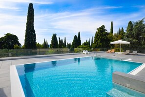 Una piscina rectangular con agua azul cristalina, rodeada por una solana con azulejos. Hay sillas de descanso y una sombrilla blanca en la solana. Se ven árboles altos y un cielo despejado al fondo.