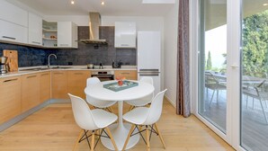 A modern kitchen with white cabinets, a round dining table, and a view of the outdoors through large sliding glass doors. - Apartment Diamante with lake view and pool (Gardone Riviera)