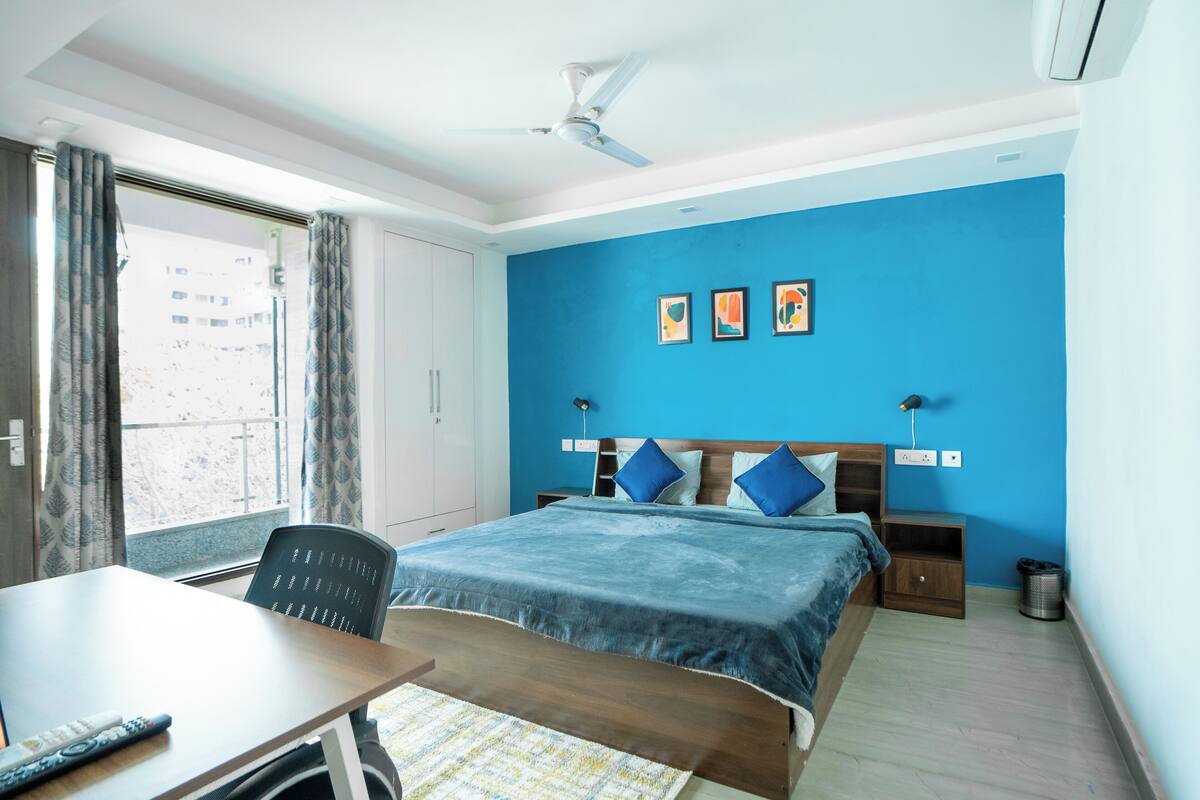 A hotel room with a bed, a desk, a chair, a window with curtains, a ceiling fan, and artwork on the wall.