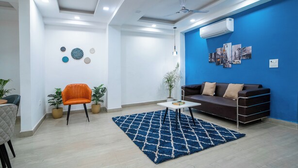 A modern living room with a blue accent wall, a brown sofa, an orange chair, a white coffee table, and a patterned rug. - FlxHo Uno - Golf Course Road (Gurugram)